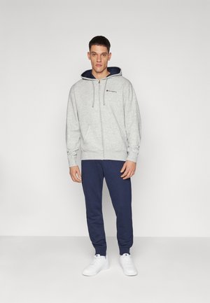 Šedá mikina na zip s námořnickou kapucí a logem, v kombinaci s námořnickými tepláky a bílými teniskami; jednobarevná paleta, ležérní design.