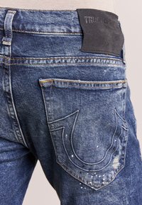 Siniset farkut, joissa on haalistunut pesu, näkyvät tikkaus, musta nahkapaikka ja koristeellinen kuvio takataskussa.