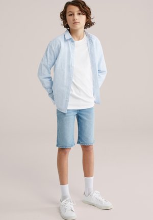 Cămașă cu dungi albastre deschis și albe, cu nasturi, purtată peste un tricou alb, asortată cu pantaloni scurți din denim deschis și pantofi sport albi.