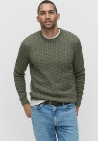 Olivgrön kabelstickad tröja med rund halsringning, strukturerat mönster och ribbad mudd, i kombination med ljusblå jeans och ett mörkt bälte.