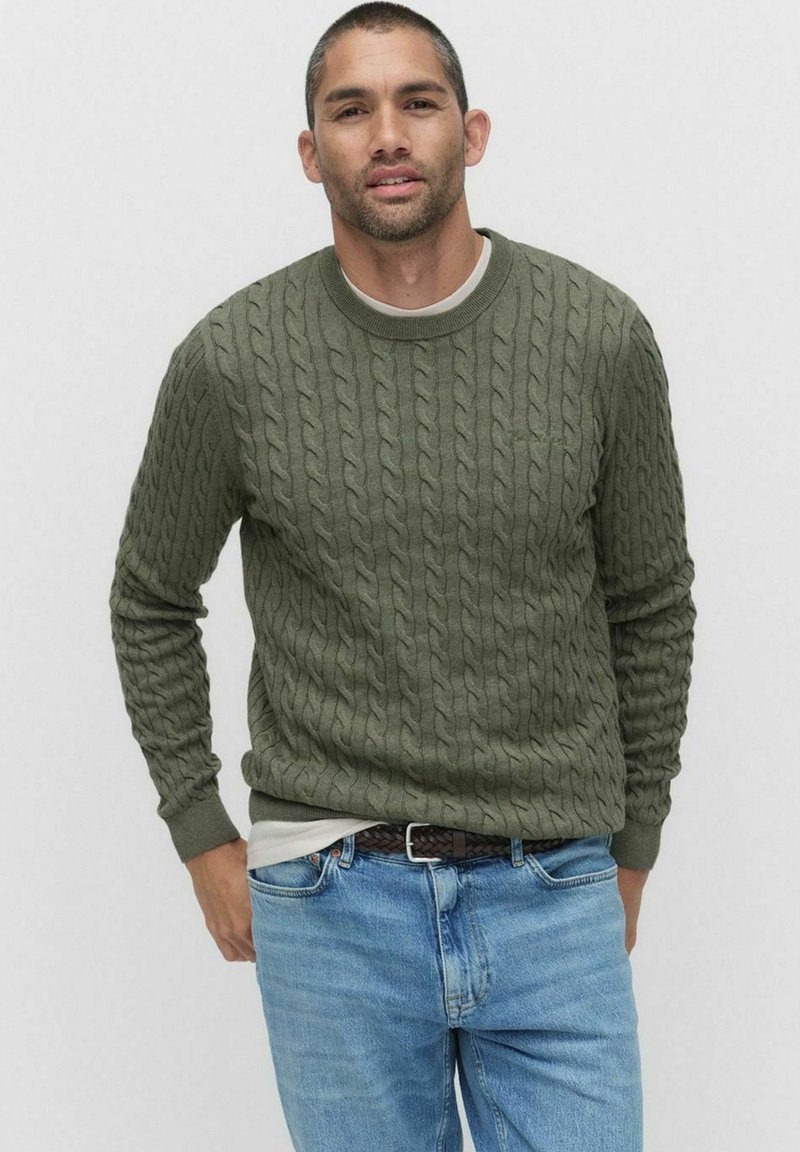 Olivgrön kabelstickad tröja med rund halsringning, strukturerat mönster och ribbad mudd, i kombination med ljusblå jeans och ett mörkt bälte.