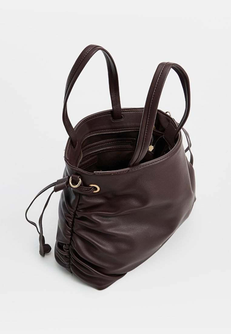 Sac fourre-tout en cuir marron avec un design doux et décontracté, fermeture à cordon, et quincaillerie en ton doré. Deux poignées de portage sont incluses.