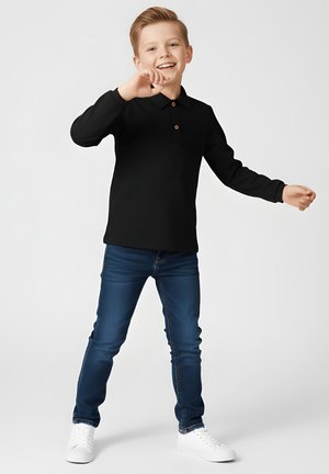 Uśmiechnięty młody chłopiec z blond włosami, ubrany w czarne, długorękawowe polo, niebieskie dżinsy i białe trampki, stojący z lekko uniesionymi ramionami.