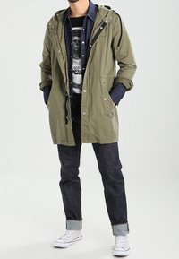 Parka vert olive avec capuche à cordon, boutons argentés et poches avant, portée sur un t-shirt graphique noir et un jean foncé.