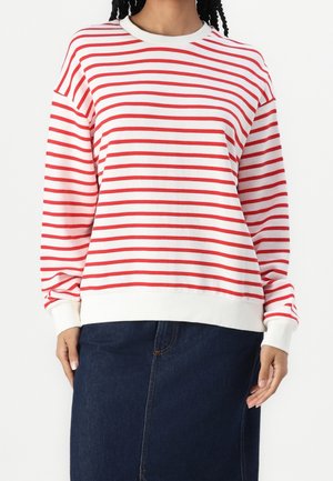 Mujer con un suéter blanco de manga larga con rayas horizontales rojas y una falda vaquera azul oscuro, de pie con las manos relajadas.