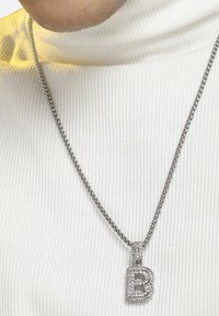Sølv kæderegnet med en vedhæng med bogstavet "B" prydet med klare, funkende rhinsten, sat mod en ribbet hvid turtleneck.