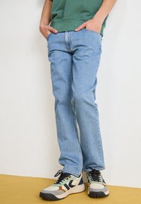 Jeans de ganga azul claro com um corte reto, apresentando um desgaste discreto e costuras em contraste. Combinados com sapatilhas atléticas multicoloridas.