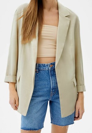 Blazer beige avec des revers en notch et des manches retroussées, associé à un débardeur léger et un short en denim bleu déchiré. Texture de tissu lisse.