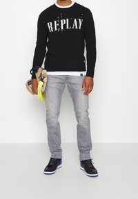 Suéter negro de punto con el logo "REPLAY", combinado con jeans gris claro. El modelo sostiene un bolso beige con una correa de neón amarilla. Zapatillas negras con acentos azules.