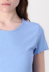 Top bleu deschis, cu fente scurte, cu margini ondulate și decolteu rotund. Materialul are o textură netedă, cu dungi verticale.