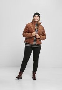 Bruine corduroy jas met een hoge kraag, zwart-grijs gestreept shirt eronder, zwarte jeans en glanzende bordeauxrode veterschoenen.