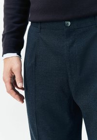 Navy textured trousers with pleats and a button fastener. Close-up view showing fabric detail and part of a hand in a dark sweater.