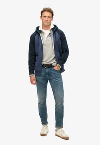 Sudadera con cremallera en color navy y gris, con bolsillos, superpuesta a una camisa polo gris, combinada con jeans azules y zapatillas blancas con detalles en color tostado.