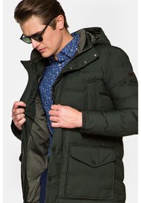 Ciemnozielona pikowana kurtka z kapturem, wyposażona w zamek błyskawiczny i zatrzask, z bocznymi kieszeniami i fakturowanym wnętrzem. Nosi się ją na niebieskiej koszuli w kwiaty.