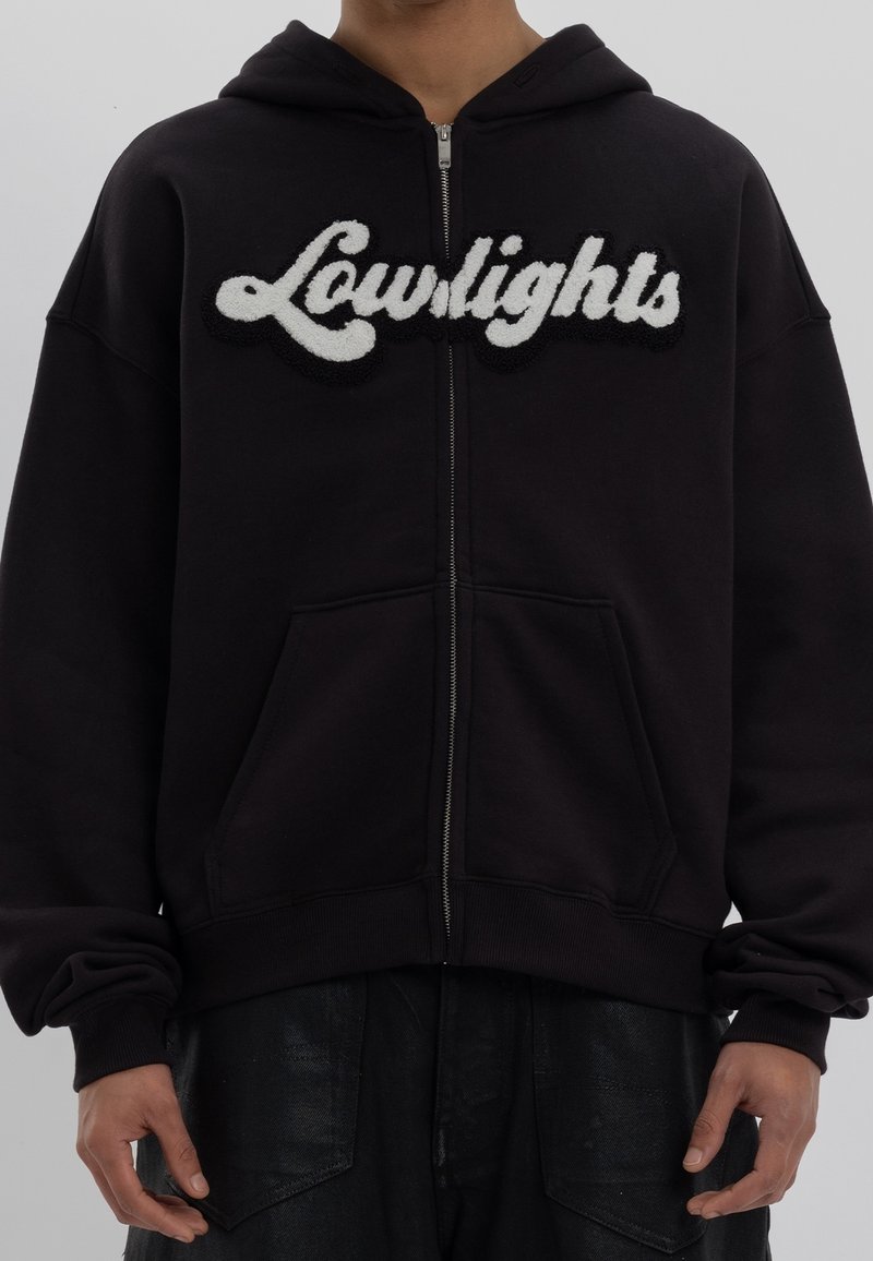 Hoodie noir à fermeture éclair avec un logo "Lowlights" blanc et duveteux. Comprend une poche avant et des poignets côtelés. Tissu doux avec une coupe décontractée.