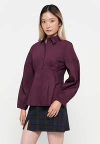 Bordeaux knoop-shirt met een gestructureerd ontwerp en lange mouwen, gecombineerd met een geruite minirok in blauw en bruintinten.