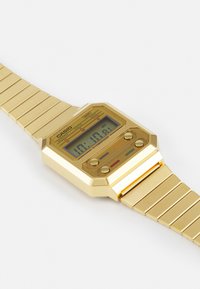 Reloj digital con caja y correa de acero inoxidable dorado. Presenta una forma octagonal, botones azul y rojo, y una pantalla digital.