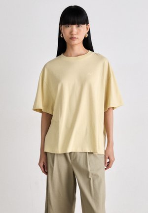 T-shirt in cotone giallo con vestibilità ampia, maniche corte e scollo rotondo, decorato con un logo discreto. Abbinato a pantaloni beige larghi.