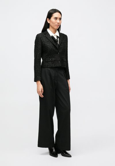 Fekete texturált dzseki, testhezálló kialakítással, két első zsebbel, fehér ing és fekete, széles szárú nadrág, valamint hegyes orrú fekete csizma fölött viselve.