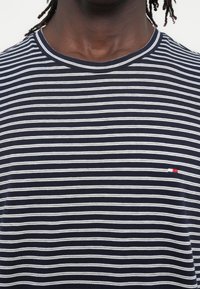 Κοντινό πλάνο σκοτεινόχρωμου ατόμου που φοράει μπλε ναυτικό και λευκή ριγέ μπλούζα με λαιμόκοψη και μικρό κόκκινο, άσπρο και μπλε εμβλημα της σημαίας στο στήθος.