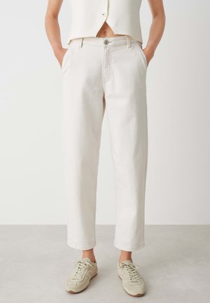 Helle beige, taillierte Jeans, mit Knopfverschluss, seitlichen Taschen und geradem Bein. Kombiniert mit grauen Sneakers.