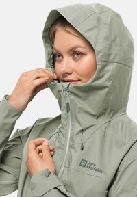 Giacca impermeabile verde chiaro con cappuccio, cerniera frontale e lacci. Presenta un logo di un verde più scuro e un tessuto liscio e testurizzato.