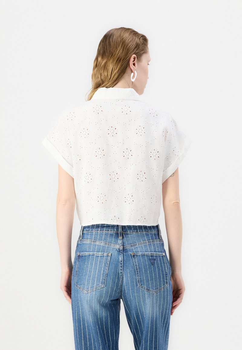 Camiseta blanca corta con patrón de ojal, mangas cortas enrolladas y cuello. Combinada con jeans azul con rayas. Se muestra la vista trasera.