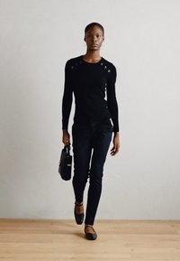 Černý dlouhý rukáv top s akcenty na ramenou, slim-fit černé kalhoty a černé baleríny. Modelka drží malou černou kabelku, zatímco kráčí.