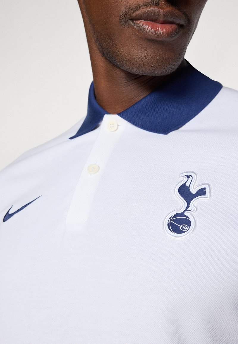 Polo shirt blanc avec un col bleu marine, présentant un logo Nike bleu marine et un écusson d'équipe brodé sur la poitrine gauche. Détail du tissu texturé.