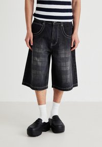 Persoană îmbrăcată în pantaloni scurți din denim cu carouri negre, șosete albe, pantofi slip-on negri și o cămașă în dungi albă și bleumarin, stând pe un podea albă.