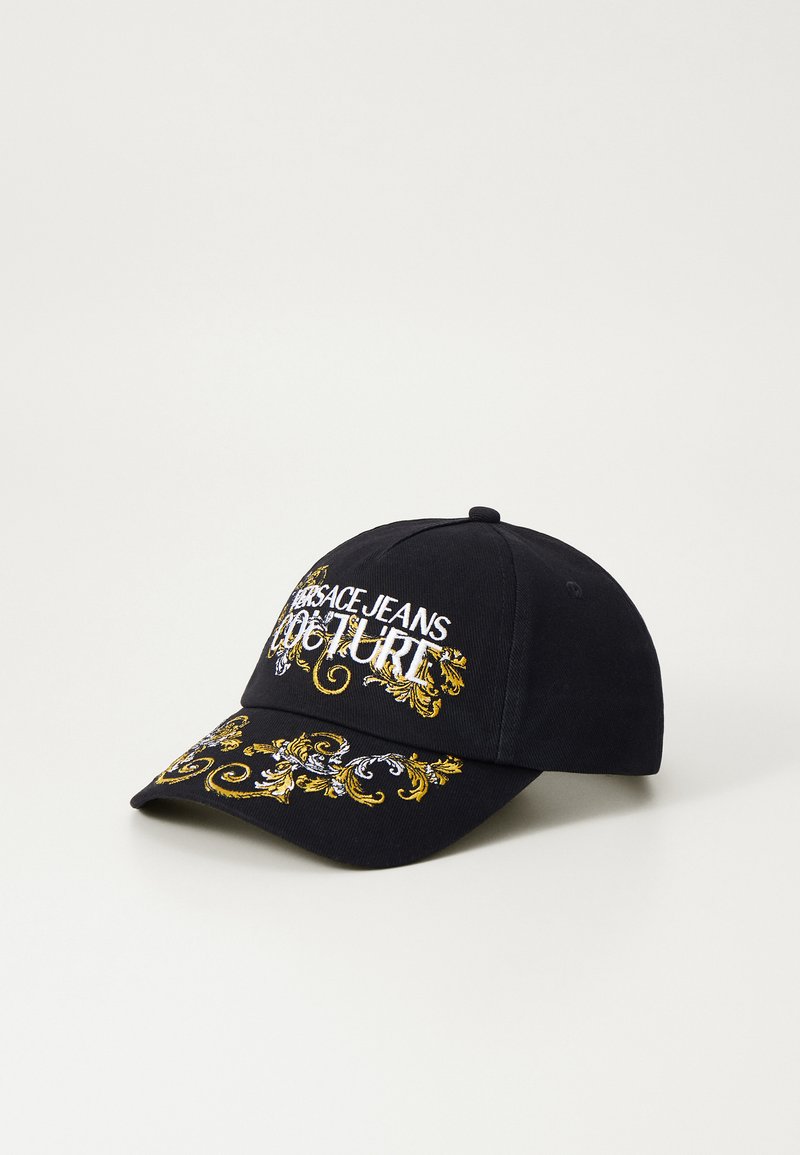 Black cap with embroidered text "VERSACE JEANS COUTURE" in white and ornate gold and silver floral designs on the visor and front.