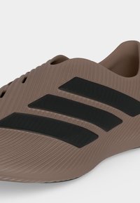 Καφέ παπούτσι τύπου slip-on με υφή στην επιφάνεια που διαθέτει τρεις μαύρες διαγώνιες ρίγες στο πλάι, χωρίς κορδόνια και με οπές αερισμού κοντά στην κορυφή.