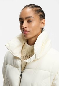 Puffjacka i ljuskräm med hög krage, med dragkedjestängning och en ribbad stickad tröja under. Slät textur och vadderad design.