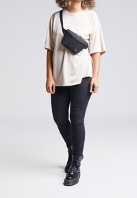 Personne portant un t-shirt oversized beige, des leggings noirs, des bottes noires à lacets et un sac bandoulière noir, se tenant contre un fond clair.