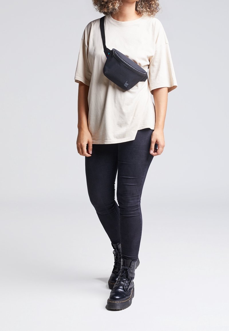 Personne portant un t-shirt oversized beige, des leggings noirs, des bottes noires à lacets et un sac bandoulière noir, se tenant contre un fond clair.