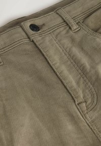 Olivengrüne Cordhosen mit Reißverschluss, einem schwarzen Knopf und schmalen vertikalen Rippungen für die Textur. Ergänzungen sind Gürtelschlaufen und Nähte.