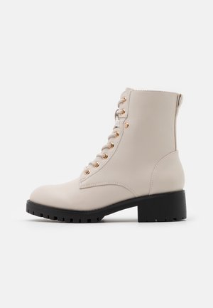 Bottine beige à lacets avec œillets dorés, semelle noire épaisse et petit talon bloc, vue de profil sur fond blanc.