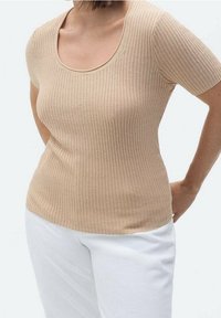 Mujer vestida con una blusa de manga corta de canalé beige y pantalones blancos, con las manos posicionadas detrás de la espalda, contra un fondo liso claro.