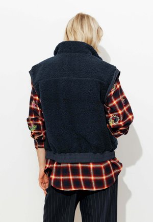 Veste en polaire bleu marine avec col montant et ourlet élastique, portée sur une chemise à carreaux rouge et noir ornée d'accents brodés colorés sur les manches.