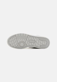 Suela de zapatilla de goma en gris claro con patrones de agarre texturizados y un detalle circular. Marca sutil incorporada en el costado.