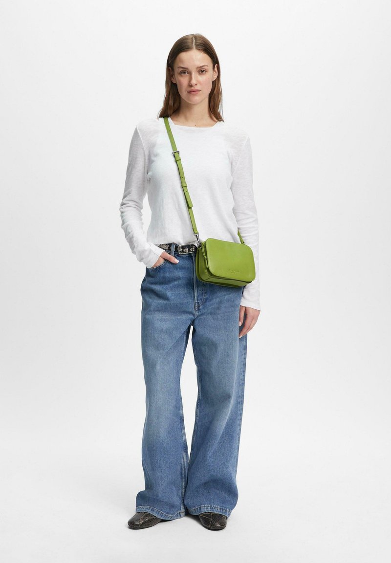 Femme portant un tee-shirt blanc à manches longues, un jean bleu ample, des chaussures noires et un sac bandoulière vert, debout devant un fond clair uni.