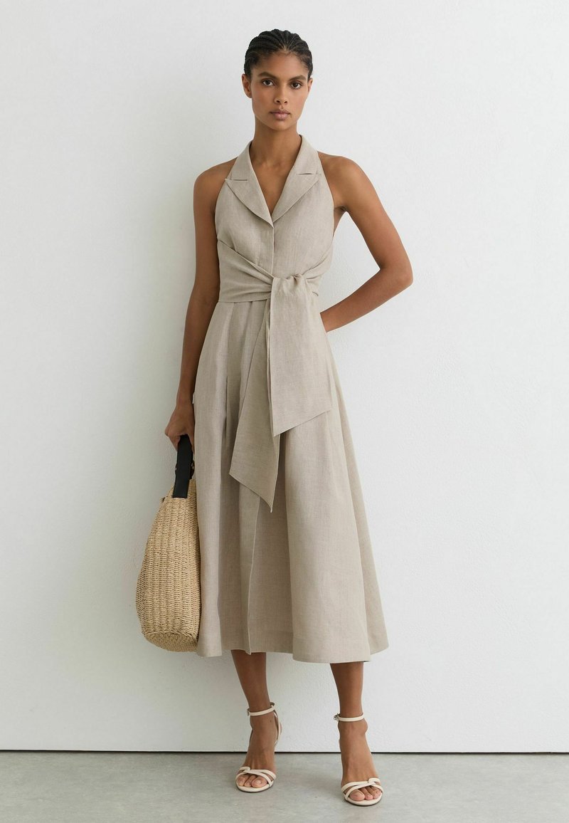 Femme portant une robe midi beige sans manches avec un grand nœud à la taille, tenant un sac à main tressé, debout contre un mur blanc avec des sandales neutres.