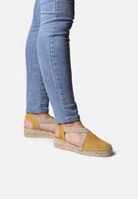 Žluté espadrilky na klínku s texturovaným svrškem a designem s křížovými pásky, umístěné na podrážce z přírodních vláken, spárované s modrými skinny džíny.