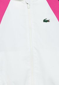 Jachetă albă cu accente roz, material texturat, închidere frontală cu fermoar și logo mic Lacoste verde. Design ușor, potrivit pentru purtarea casual.