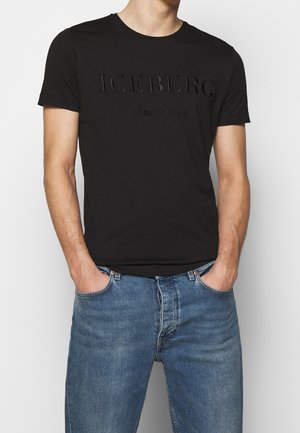 Homme portant un t-shirt noir à manches courtes avec le texte "ICEBERG depuis 1974" et un jean bleu en denim, mains dans les poches, cadré au niveau du cou et des cuisses.