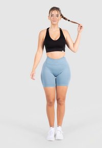 Zwarte sportbeha en lichtblauwe high-waist shorts. Model staat met een paardenstaart, draagt witte sneakers, op een neutrale achtergrond.