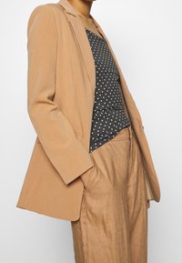 Beige blazer van zacht materiaal met een getailleerde pasvorm, gedragen over een grijze tanktop met stippen, gecombineerd met losse beige broeken.