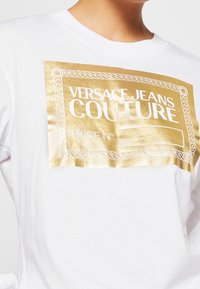 Bílý tričko s velkým kovovým zlatým obdélníkovým logem s vyraženým textem a okrajovým designem ve tvaru lana. Krátké rukávy a kulatý výstřih.