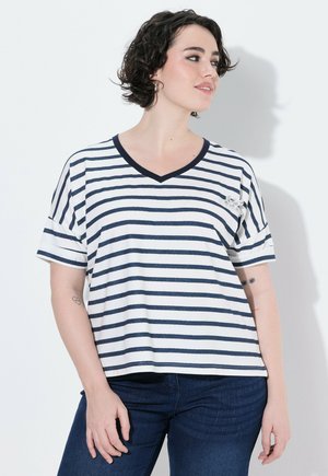 Kvinde med kort krøllet hår iført en hvid og marineblå stribet V-udskåret skjorte og mørkeblå jeans, kigger til siden mod en hvid baggrund.