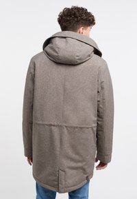 Brun texturerad kappa med huva, med en sömnadslogotyp på baksidan och rak skärning. Bärs över blå jeans.